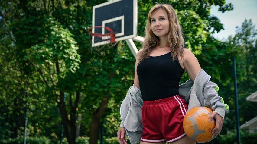 Confident woman posing with a basketball on an outdoor court surrounded by lush greenery.