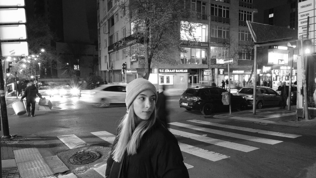 A woman in a beanie and jacket stands at a city crosswalk at night in an urban setting.